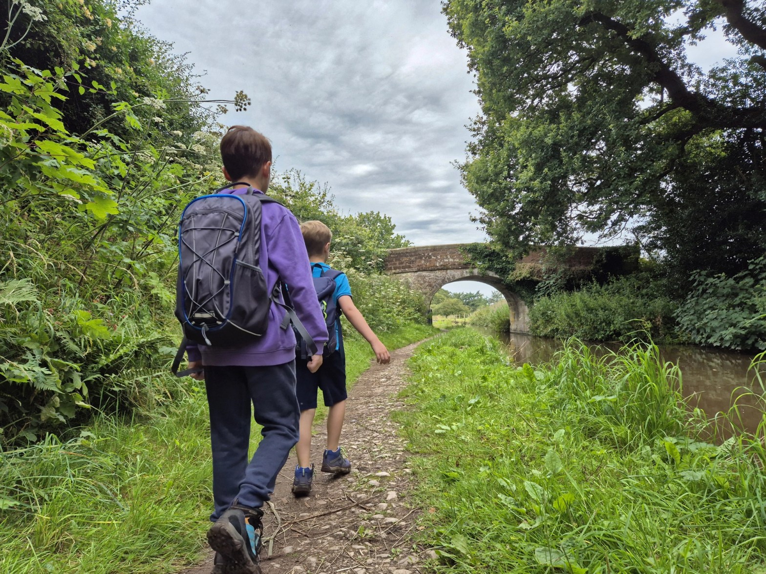 2 boys with backpacks walk alongside the Shropshire union Canal and are approaching a bridge.