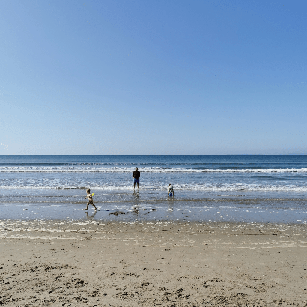 2 children, supervised by their dad, run around in the beach as the sea gently laps their feet, with a bucket and spade