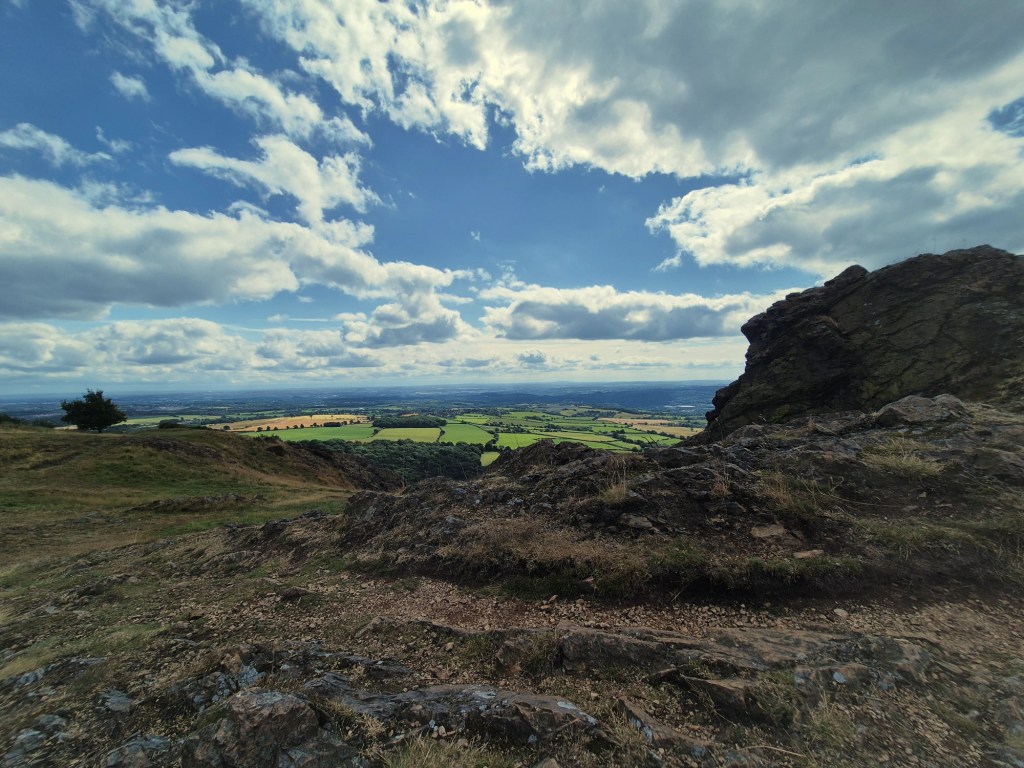 A rocky foreground leads to blue skies and green fields in the background