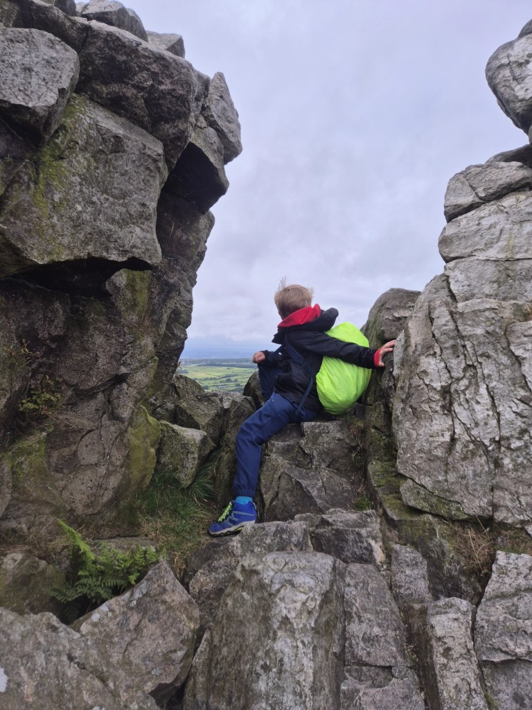 a child in wet weather gear and a backpack sits between two large rock formations looking towards the view on the other side