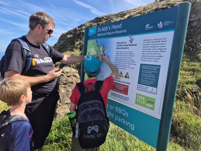 2 boys and a man look at and point at a board with a map of the coastal walk on