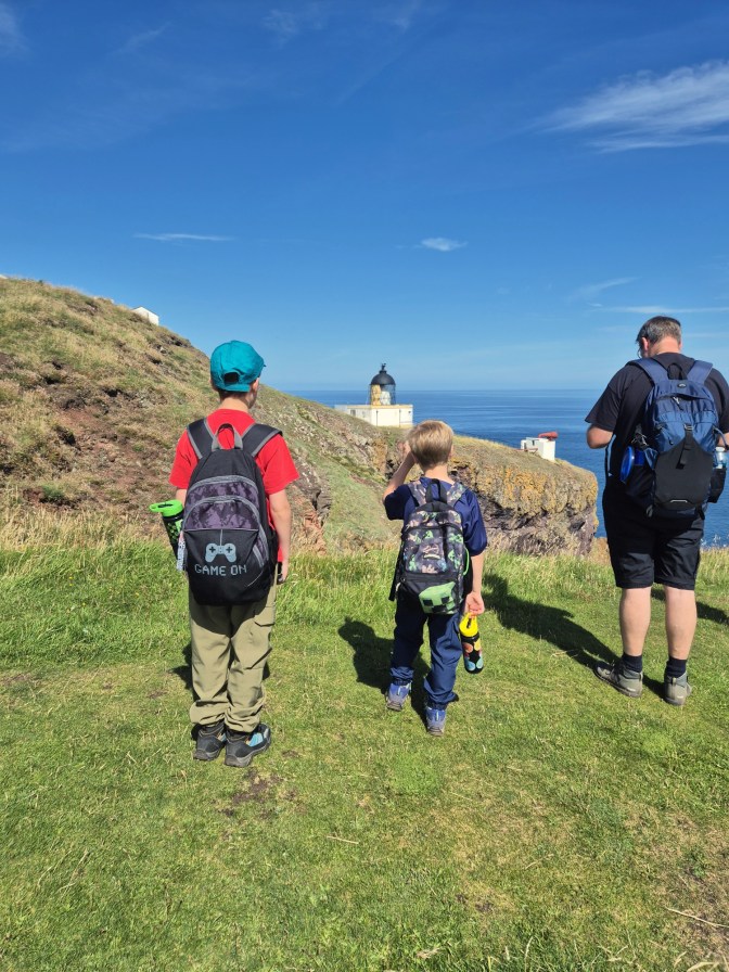 2 young boys and a man, all with backpacks, stand on a cliff edge looking towards a lighthouse
