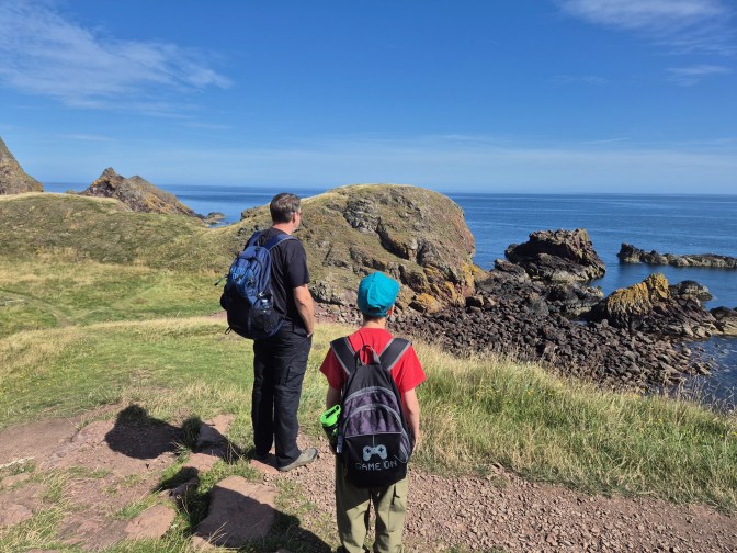 A boy and a man with backpacks walk across grassland with the sea and skies in the background