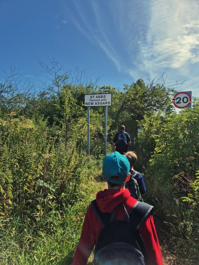 2 boys with backpacks walk through a green pathway with a sign saying ST ABBS TWINNED WITH NEW ASGARD