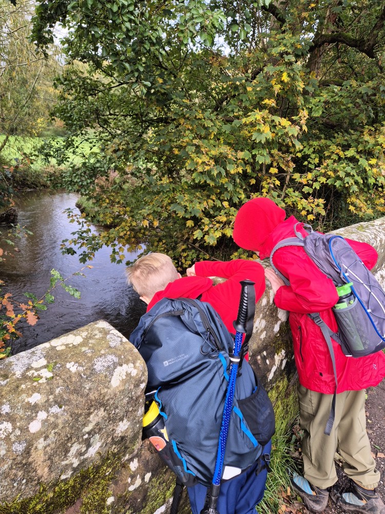 Two boys peer over a bridge into a canal. They are both wearing outdoor gear and backpacks
