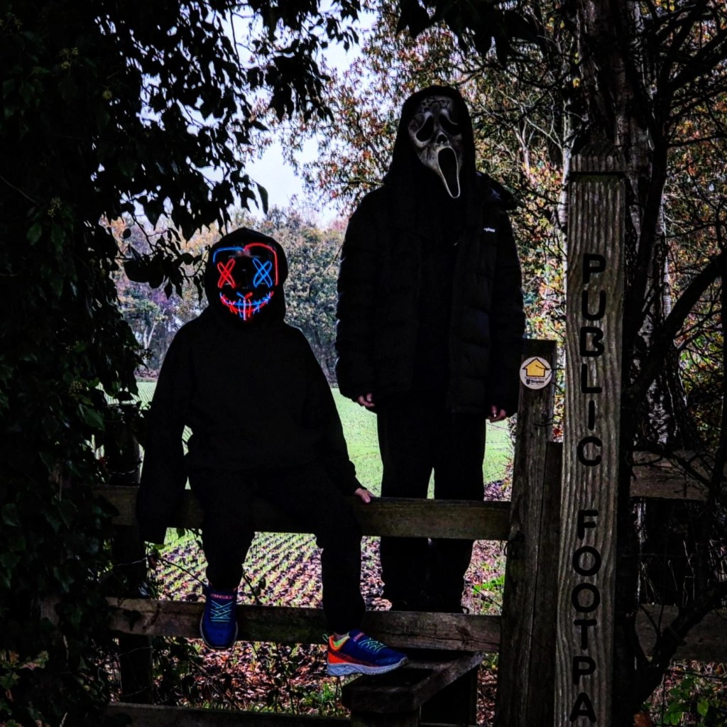 2 boys dressed in black with halloween maskes go for a halloween walk and pose on a stile