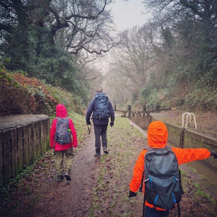 2 boys and a man walk down a canal on a grim day. the two children are wearing bright orange coats and backpacks, the man is wearing a dark coloured coat and a backpack