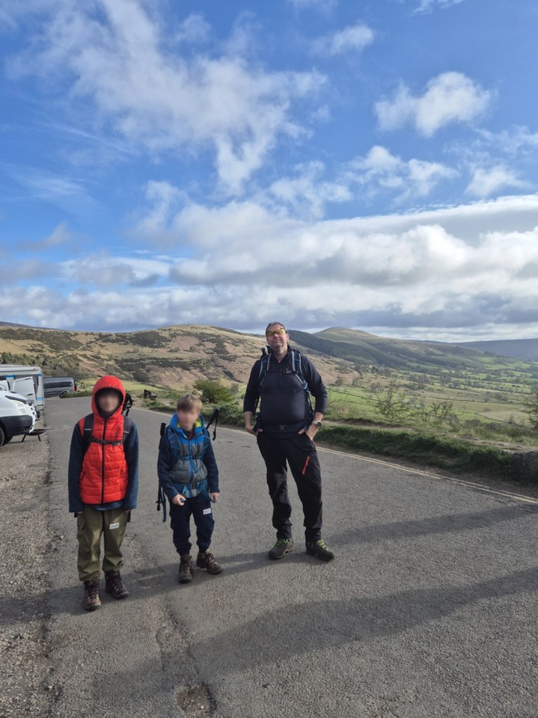 A man and two children with walking gear stand with rolling hills in the distance.