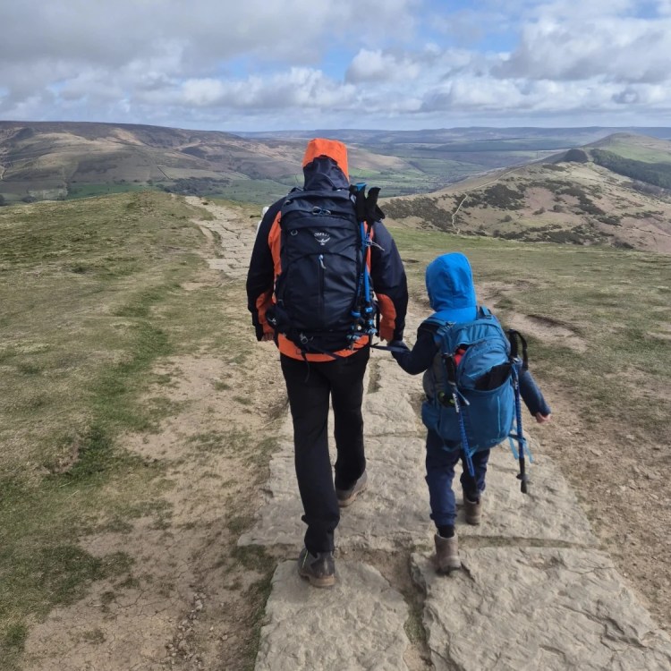 a boy and a man walk along a paved path on the ridge of a hill mam tor wearing outdoor gear and backpacks