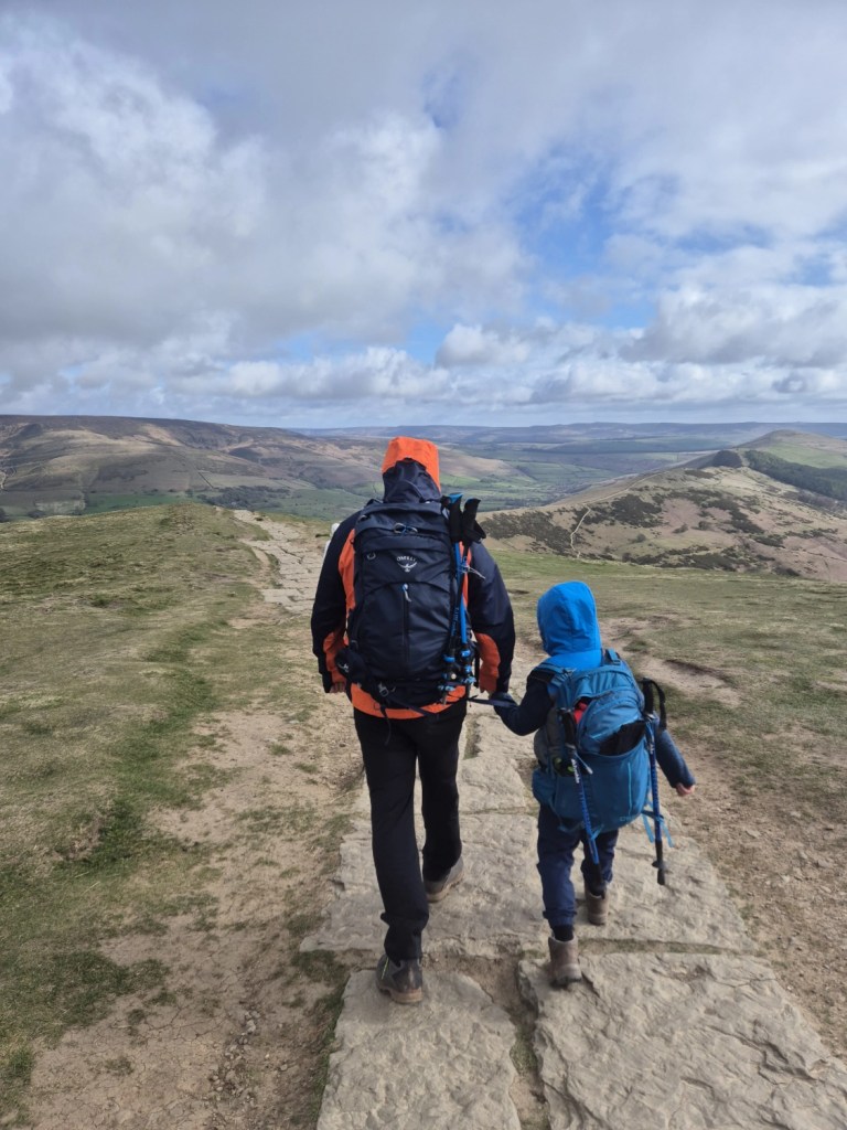A man and a child walk along a narrow ridge with hills in the distance