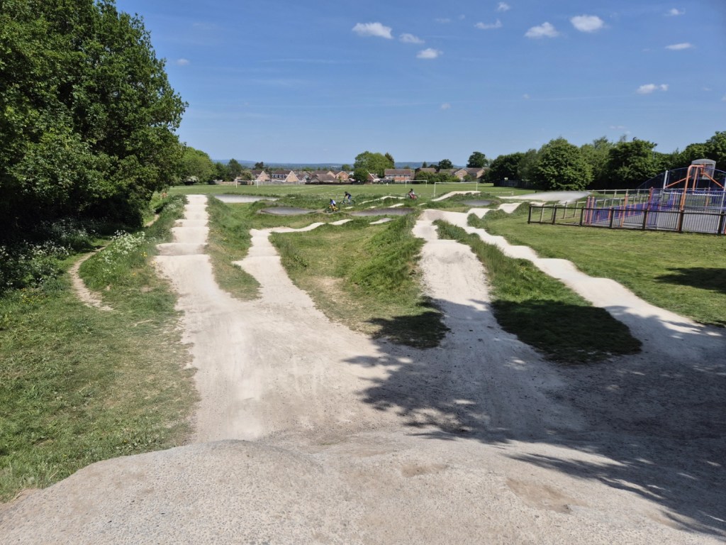 A wide view of Bayston Hill Pump Track on a sunny day, featuring multiple dirt biking lanes with grassy edges, surrounded by trees and a nearby children’s playground. A few riders are visible in the distance.