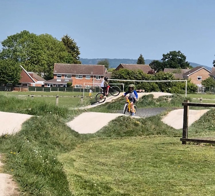 Two children riding bikes on a pump track; one performs a wheelie while the other rides over a small bump. The track is surrounded by grass, with houses and hills in the background under a clear blue sky.