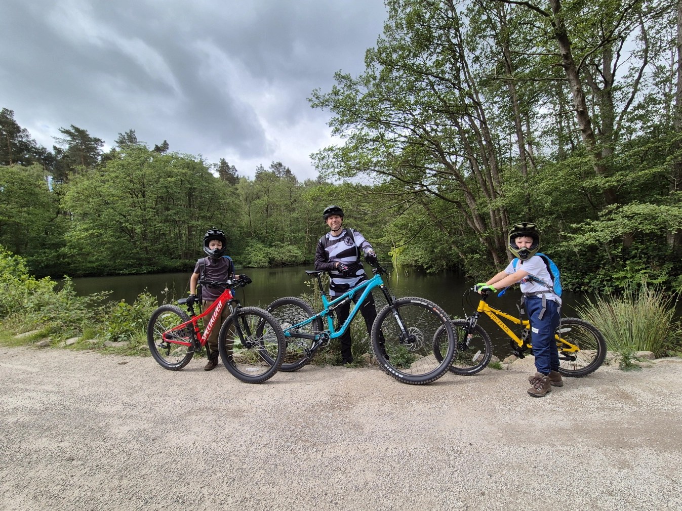 Two children and an adult pose with their mountain bikes beside a woodland lake at Cannock Chase, all wearing helmets and biking gear, ready for a family trail ride.