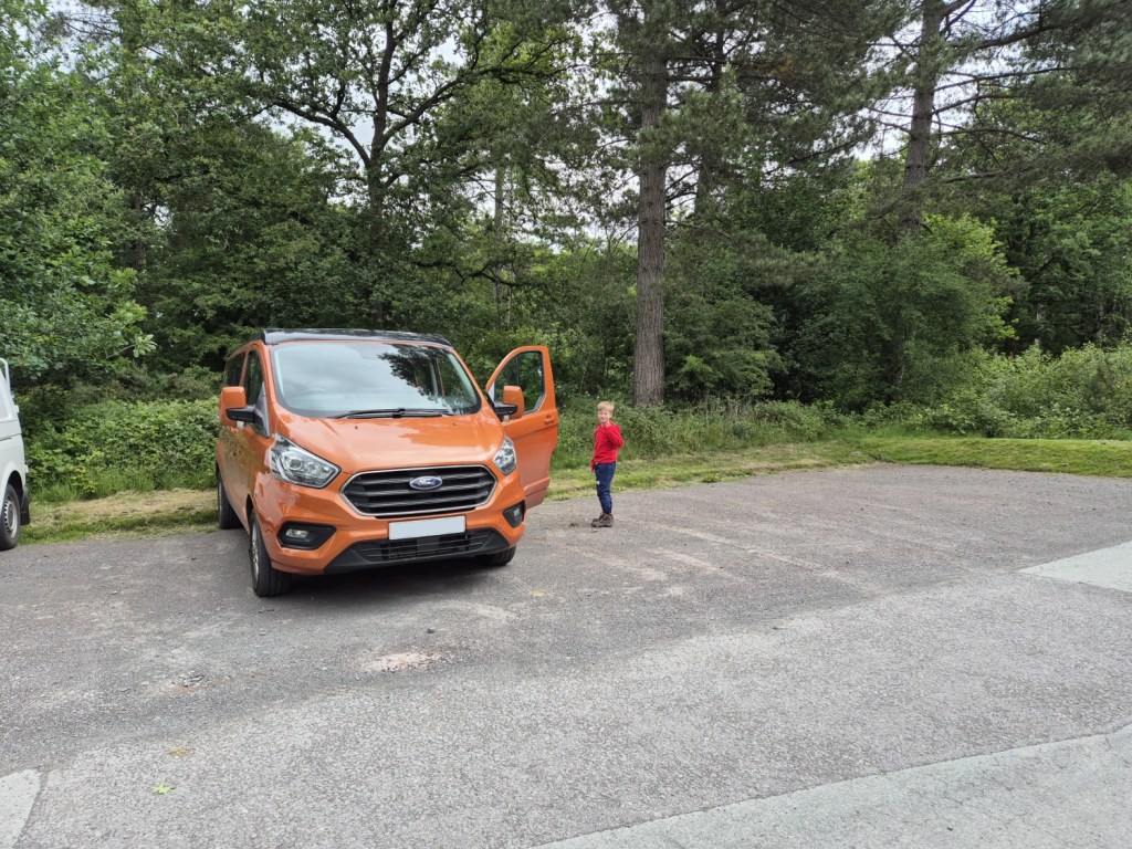 Young boy standing beside an orange Ford Transit Custom campervan in the Haughmond Hill car park, surrounded by tall pine trees and greenery.
