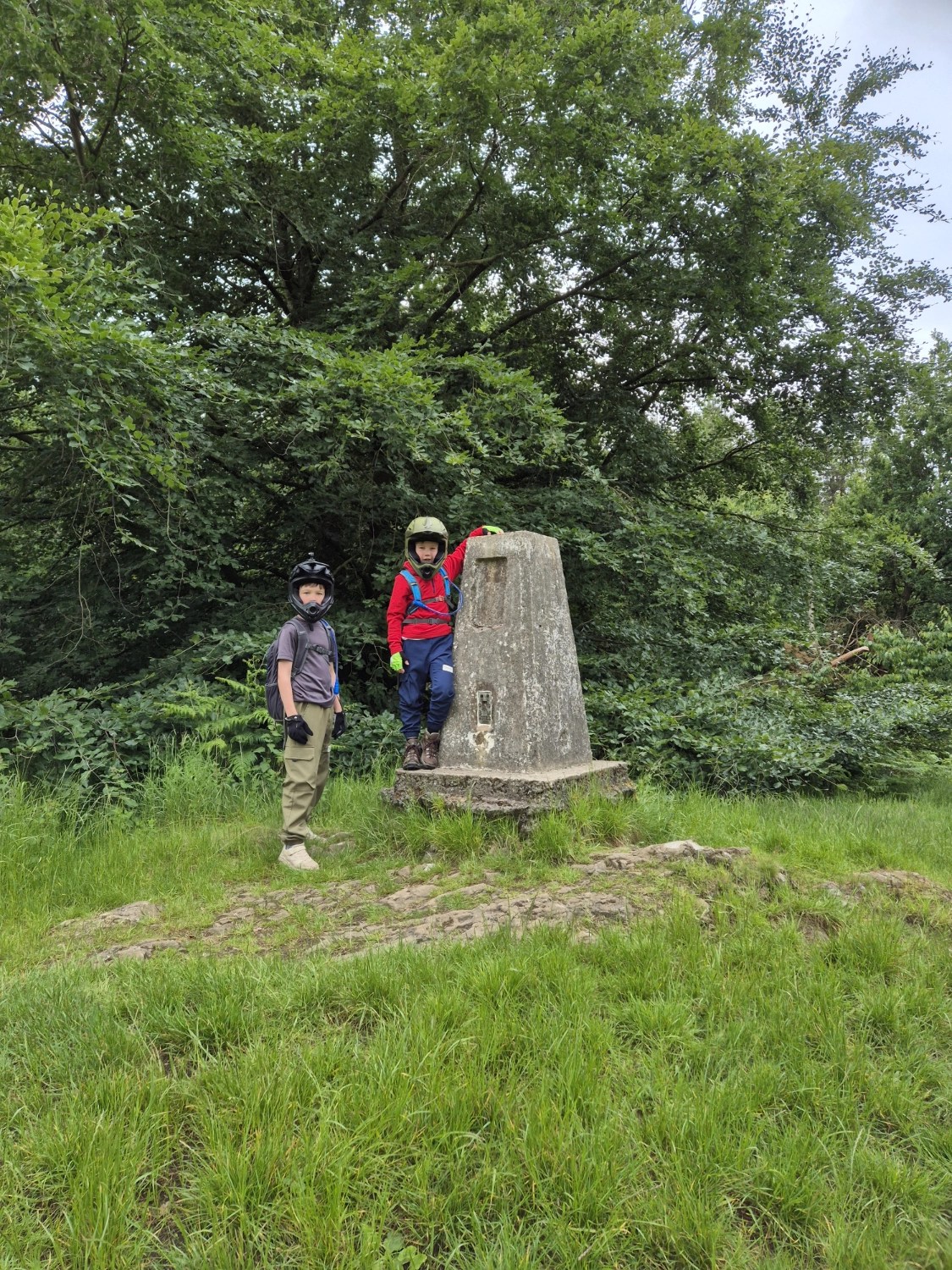 Two children in biking gear standing beside the concrete trig point marker at Haughmond Hill, surrounded by woodland.