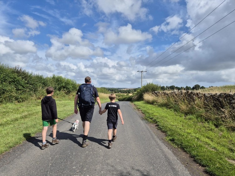 2 boys are walking along a country road, one boy is holding the lead to a jack russel and the other boy is holding the mans hand, the man is wearing a backpack