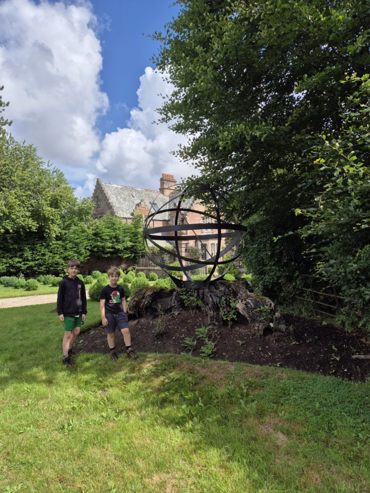 2 boys stand on grass near a flower bed and an orb like sculpture