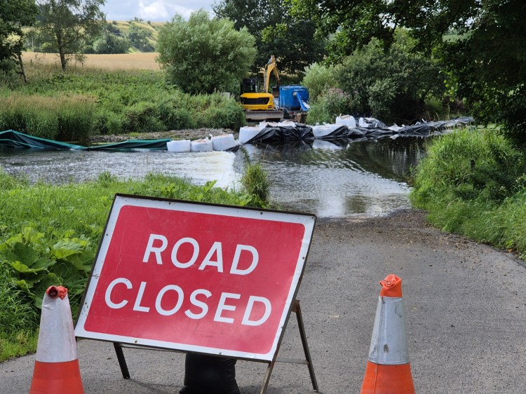 a road closure sign stands in front of a river with a ford. beyong that and halfway up the ford are a dozen tonne bags and a small digger blocking the way
