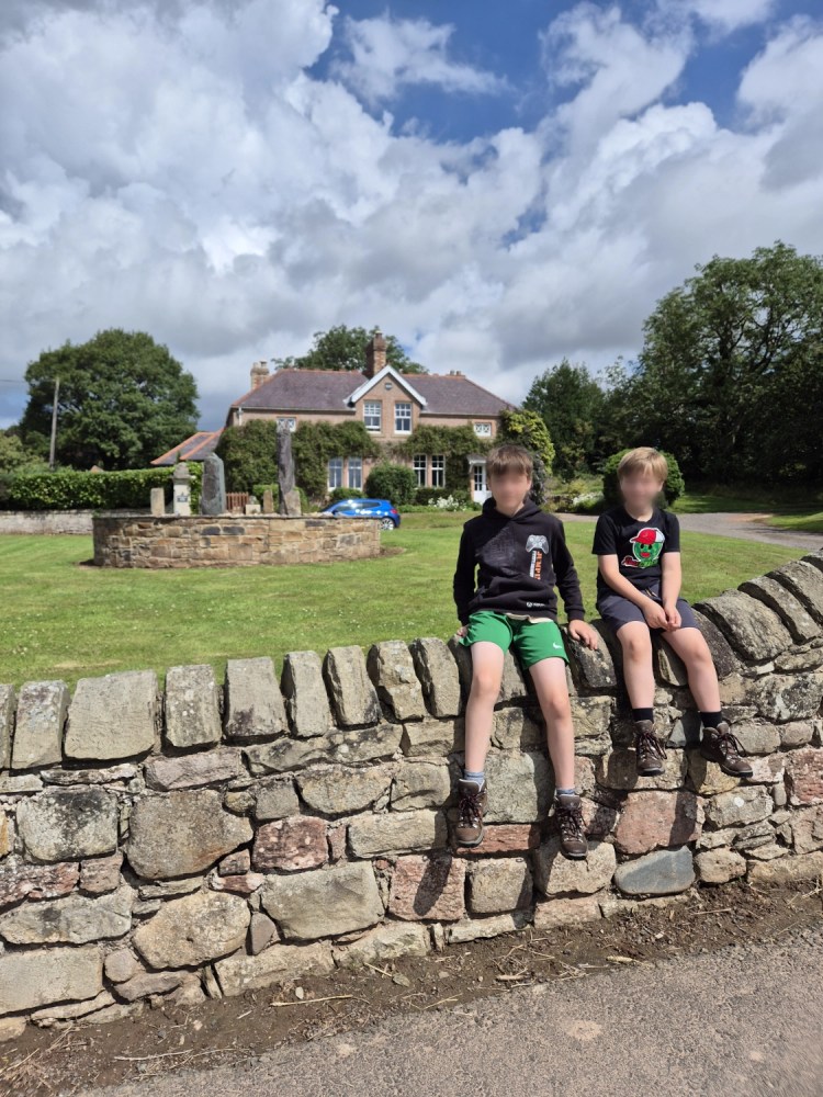 2 boys sit on an old stone wall with a house in the background. in the garden is a round flower bed with a standing stones structure within it.