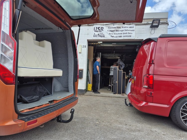 View of campervan parked outside a conversion workshop with the back doors open.