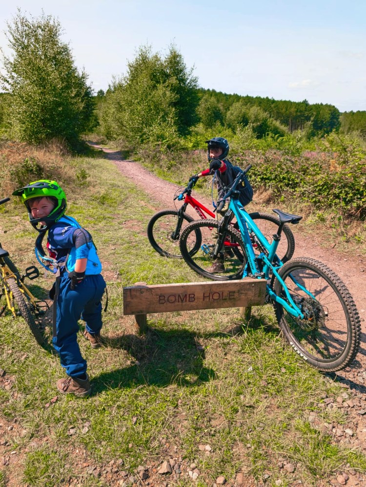 Two children with mountain bikes smiling beside a small wooden jump marked “BOMB HOLE” on a forest trail.