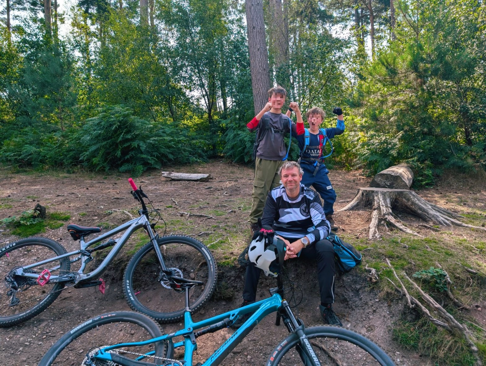 Family taking a break on a forest trail, sitting near their mountain bikes, smiling and cheering.