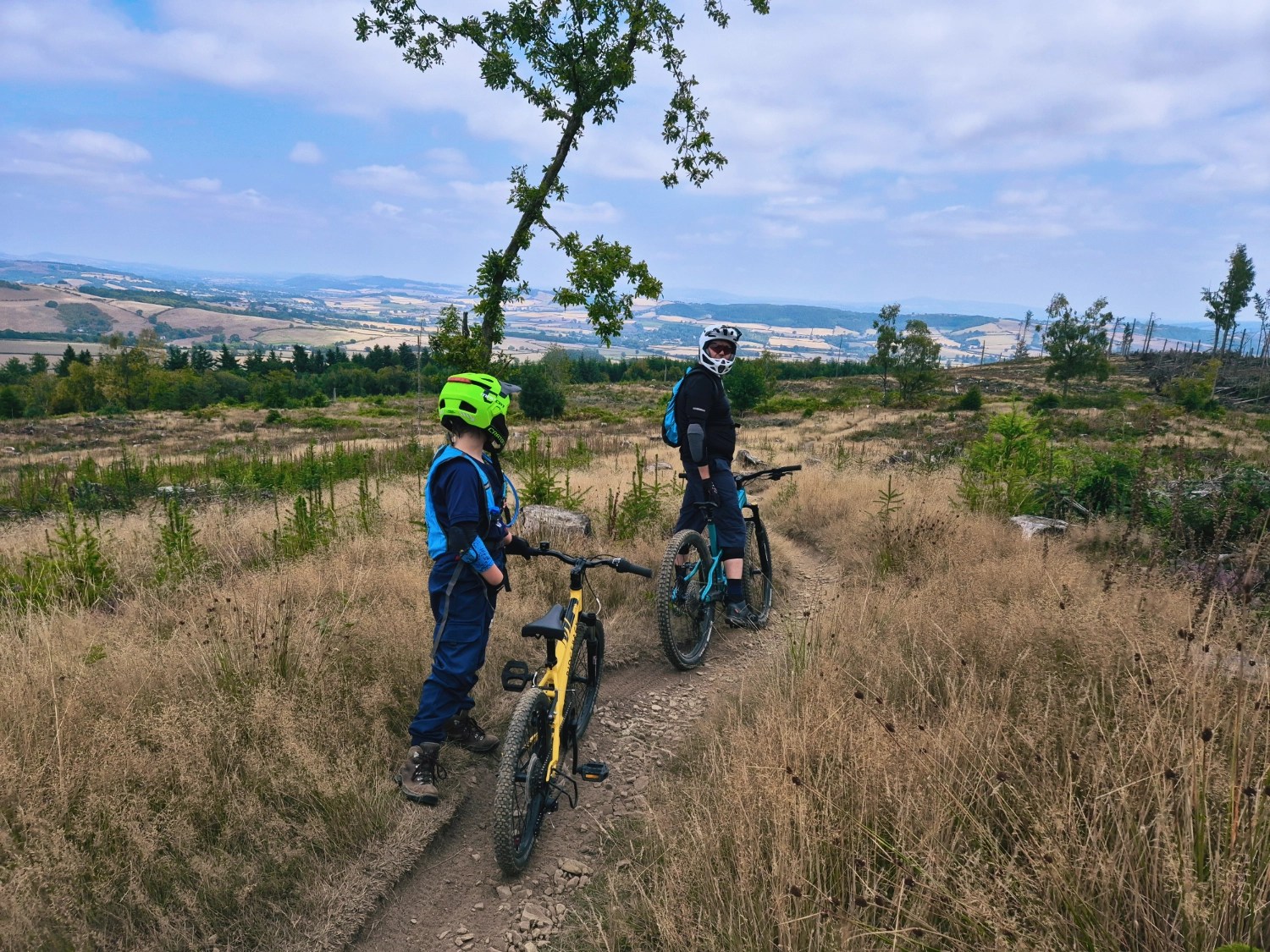 Child and Dad with mountain bikes on a grassy, gravelly trail overlooking wide countryside views.