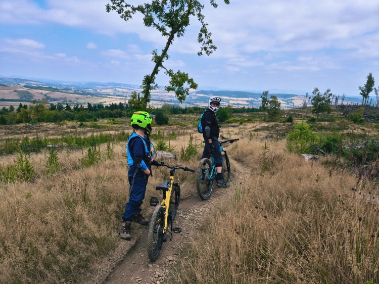 Child and Dad with mountain bikes on a grassy, gravelly trail overlooking wide countryside views.