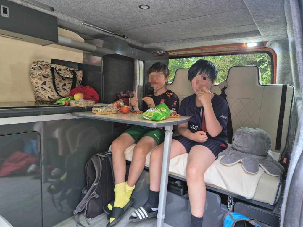 Two children eating at a table inside a campervan, with food and drinks on the counter.