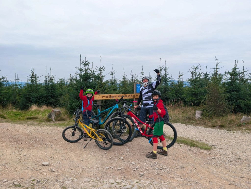 Two children and an adult with mountain bikes, standing on a gravel trail surrounded by trees.