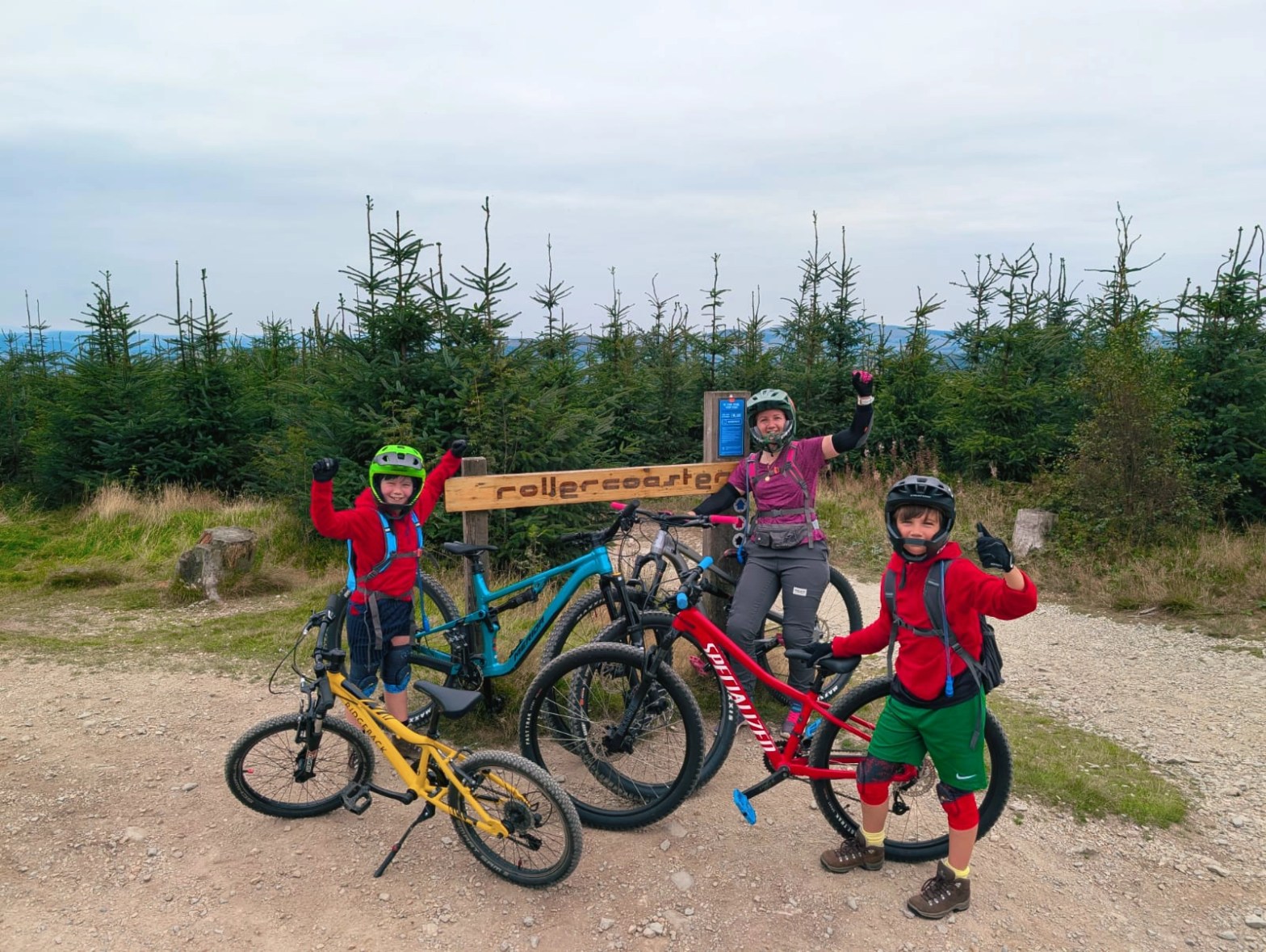 Two children and an adult with mountain bikes, standing on a gravel trail surrounded by trees.