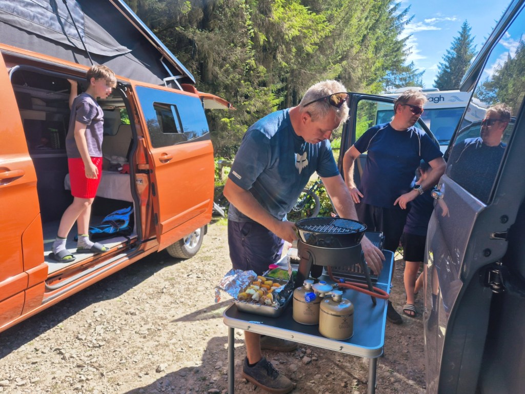 2 men between 2 campervans cooking with a child in the door of the orange van