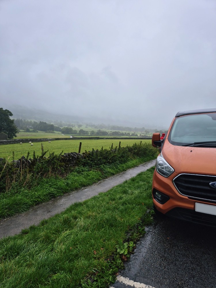 Orange campervan parked on a rainy Peak District lane with misty hills in the background.