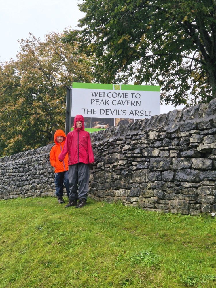 Two children in bright waterproofs standing by the “Welcome to the Peak Cavern – The Devil’s Arse” sign in Castleton.