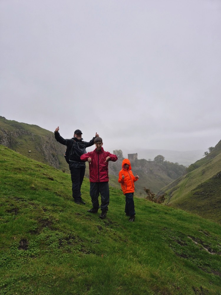 Two children standing on a grassy slope, arms raised in excitement, with an adult behind them in the Peak District rain.