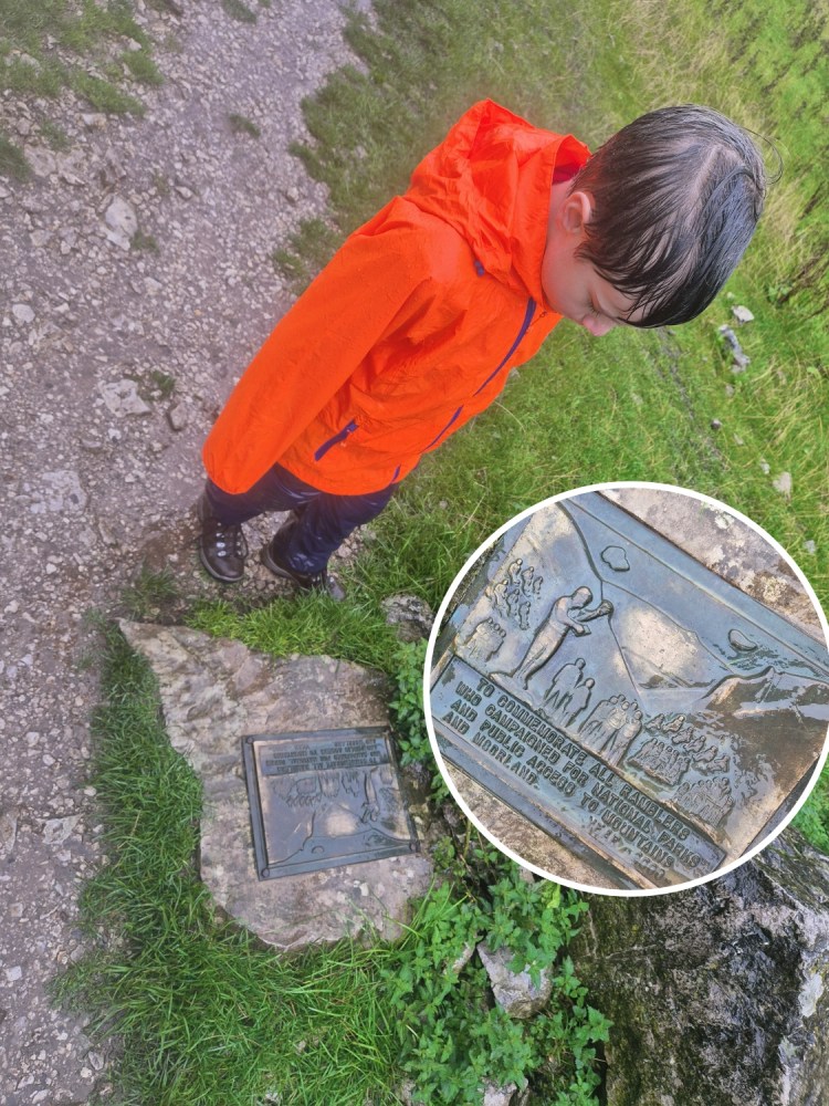 Child in an orange coat looking down at a wet stone waymarker disc with place names engraved.